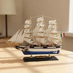 Italian Confection tall ship wooden model displayed on a light wood desk in a warm home setting, showing three-mast full-rigged white sails, blue-and-white striped hull, Italian flag, and brass Confection nameplate on black display base