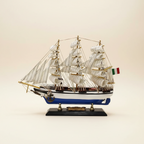 Side profile of Italian Confection tall ship wooden model on cream background, showing complete three-mast silhouette with full white sails, blue-and-white striped hull, Italian flag at stern, anchor detail, and black display base with brass nameplate