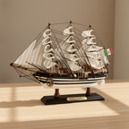 Amerigo Vespucci Italian Navy tall ship wooden model displayed on a wooden desk in a warm home setting, showing three-mast full-rigged white sails, black-and-white striped hull with white dot trim, dense black rope rigging, Italian Navy flag, and black display base with gold AMERIGO VESPUCCI nameplate