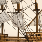 Close-up detail of Mayflower wooden ship model showing white striped sail fabric texture, dark rope rigging knots tied to wooden yard arms, three-mast cross-beam structure, and warm walnut-tone deck planking craftsmanship

