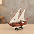 Handcrafted Kuwait Dhow wooden ship model displayed on a warm wooden desk in a home setting, white lateen sails, mahogany-toned hull, authentic rope rigging, black display stand with custom engraved nameplate, soft warm natural light
