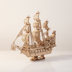 Kit de maqueta de madera Queen Anne's Revenge en vista frontal de tres cuartos, con tres mástiles y velas curvas, decoración de calaveras en el casco, bandera pirata en el bauprés, popa ornamentada y base con placa de nombre sobre fondo blanco crema