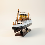 Modelo de madera del RMS Titanic en vista frontal de tres cuartos, mostrando proa con detalle de ancla y letras TITANIC, cuatro chimeneas amarillas con tapas negras, dos mástiles de madera con jarcia negra de alambre, barandillas blancas de cubierta superior y base de exhibición de madera oscura con placa TITANIC sobre fondo blanco crema