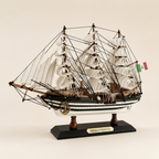 Side profile of Amerigo Vespucci Italian Navy tall ship wooden model on cream background, showing complete three-mast silhouette with full white sails, dense black rope rigging, black-and-white striped hull with white dot trim, Italian Navy flag, anchor at bow, and black display base with gold nameplate