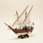 Handcrafted Kuwait Dhow wooden ship model front three-quarter view showing white lateen sails, mahogany hull planking, rope rigging, and black display stand with engraved nameplate, cream white background