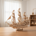 Kit de maqueta de madera Queen Anne's Revenge sobre un escritorio de roble claro en un ambiente cálido del hogar, barco pirata de tres mástiles con casco decorado con calaveras, velas curvas y popa ornamentada bajo luz natural suave