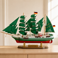 Alexander von Humboldt green-sail wooden tall ship model displayed on a light wood cabinet in a bright modern interior, featuring emerald fabric sails, German flags, detailed rigging, green and white hull, red lower hull and polished wooden display base.