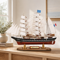 Belem wooden tall ship model displayed on a light wood cabinet in a bright modern interior, featuring white sails, black and red hull, French flags, detailed rigging and a polished wooden base.