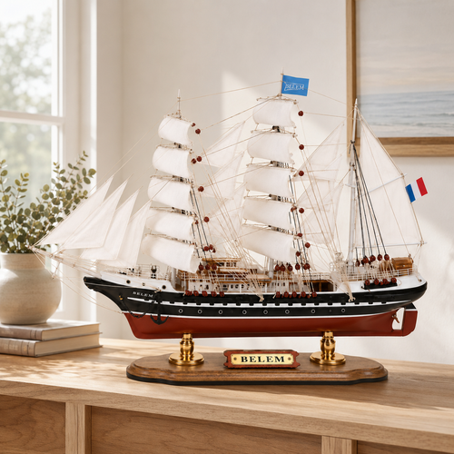 Belem wooden tall ship model displayed on a light wood cabinet in a bright modern interior, featuring white sails, black and red hull, French flags, detailed rigging and a polished wooden base.