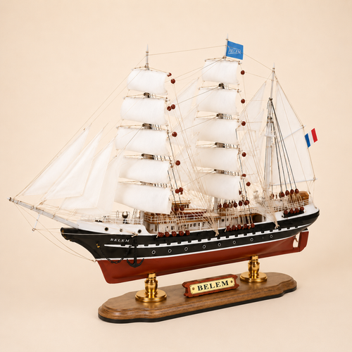 Front 45-degree view of the Belem wooden sailing ship model on a cream background, showing the bow, layered white fabric sails, black hull, red lower hull and French tricolor flag.