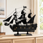 Black Pearl wooden pirate ship model displayed on a light wood cabinet in a bright modern interior, featuring black fabric sails, skull-and-crossbones graphics, detailed rigging, black hull, gold accents and a black display base.