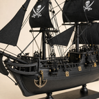 Close-up of the Black Pearl model ship showing textured black sails, skull-and-crossbones graphics, fine rope rigging, pulleys, deck railings, anchor, gold fittings and handcrafted hull details.