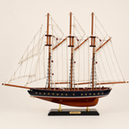 Atlantic handcrafted wooden sailboat model full side profile view on cream white background, three tall wooden masts with white sails and intricate rope rigging, black hull with gold porthole row and warm walnut-tone keel