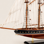 Close-up detail of Atlantic wooden ship model showing handcrafted rope rigging, white canvas sails tied to wooden yards, brass porthole fittings along black lacquered hull, and fine wood grain craftsmanship