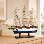 Italian Confection wooden tall ship model displayed on a light wood cabinet in a bright modern interior, featuring cream fabric sails, gold-tone masts, detailed rigging, blue and white hull, anchor and black display base.