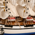 Close-up of the Italian Confection model ship showing textured cream sails, fine rope rigging, black pulleys, wooden deck, red-roofed cabins, white railings and handcrafted nautical details.