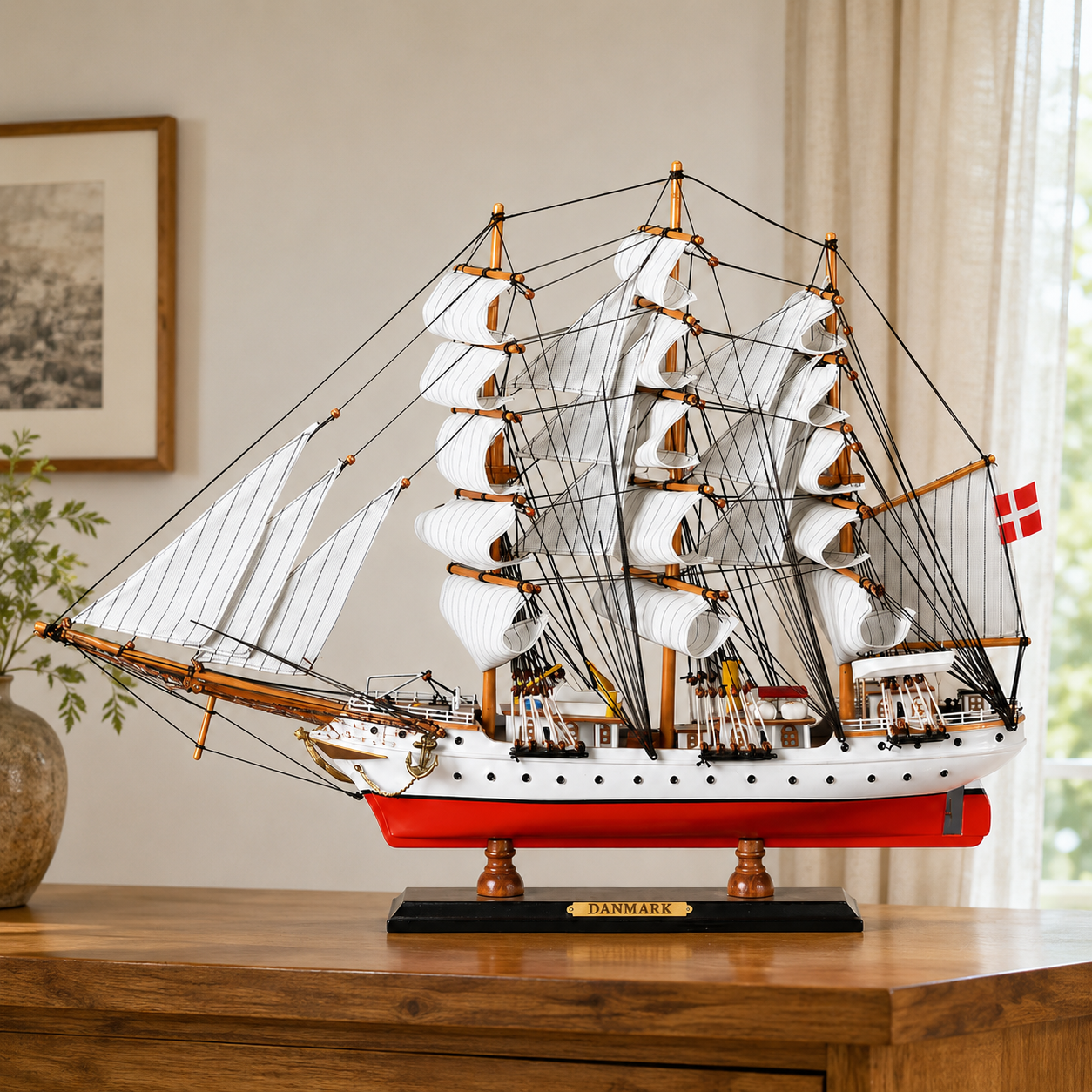 Large Danmark wooden tall ship model displayed on a wooden table, featuring white fabric sails, detailed black rigging, a white and red hull, Danish flag and black display base.