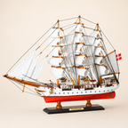 Front 45-degree view of the Danmark sailing ship model on a cream background, showing the bow, layered white sails, wooden masts, intricate rigging and Danish flag.