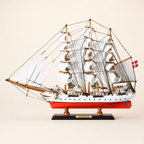 Side view of the Danmark three-masted tall ship model featuring white fabric sails, dense black rigging, a white hull, bright red lower hull and black presentation stand.