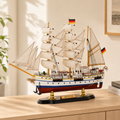 Gorch Fock wooden tall ship model displayed on a light wood cabinet in a bright modern interior, featuring cream fabric sails, detailed rigging, German flags, white hull with blue and gold accents, red lower hull and black display base.