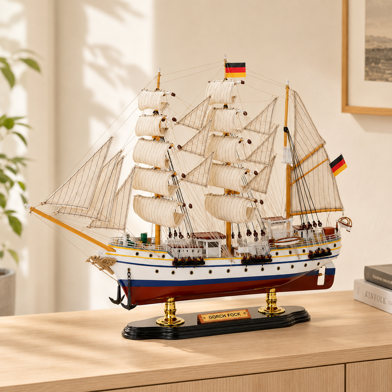 Gorch Fock wooden tall ship model displayed on a light wood cabinet in a bright modern interior, featuring cream fabric sails, detailed rigging, German flags, white hull with blue and gold accents, red lower hull and black display base.