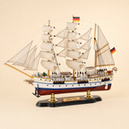 Front 45-degree view of the Gorch Fock wooden sailing ship model on a cream background, showing the bow, layered cream sails, gold-tone masts, black anchors, German flags and polished display stand.