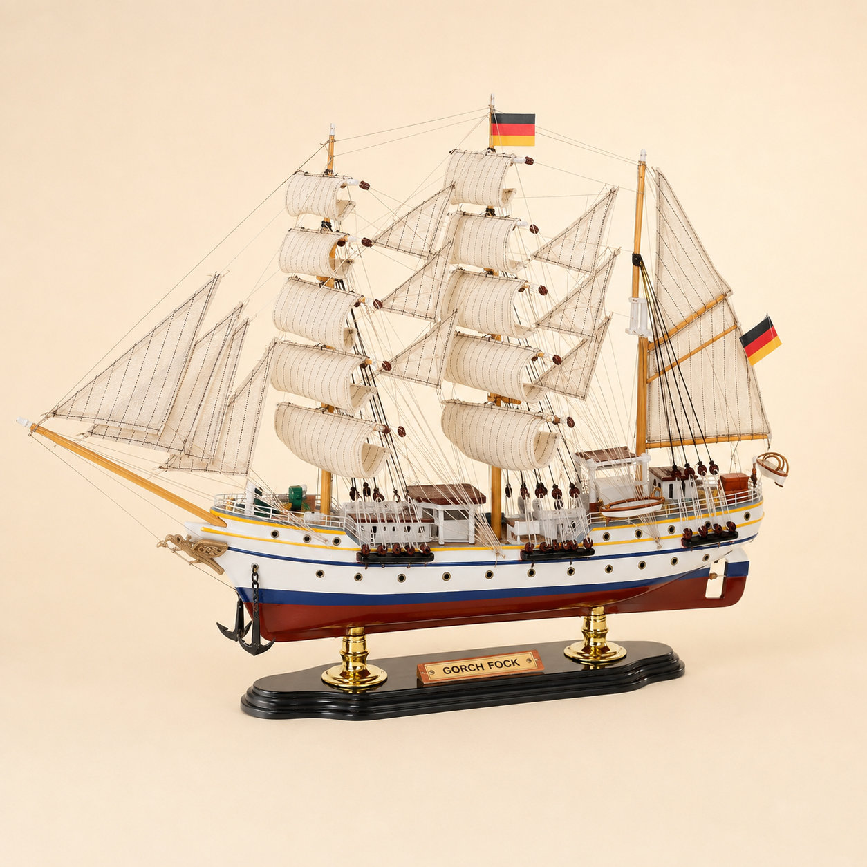Front 45-degree view of the Gorch Fock wooden sailing ship model on a cream background, showing the bow, layered cream sails, gold-tone masts, black anchors, German flags and polished display stand.