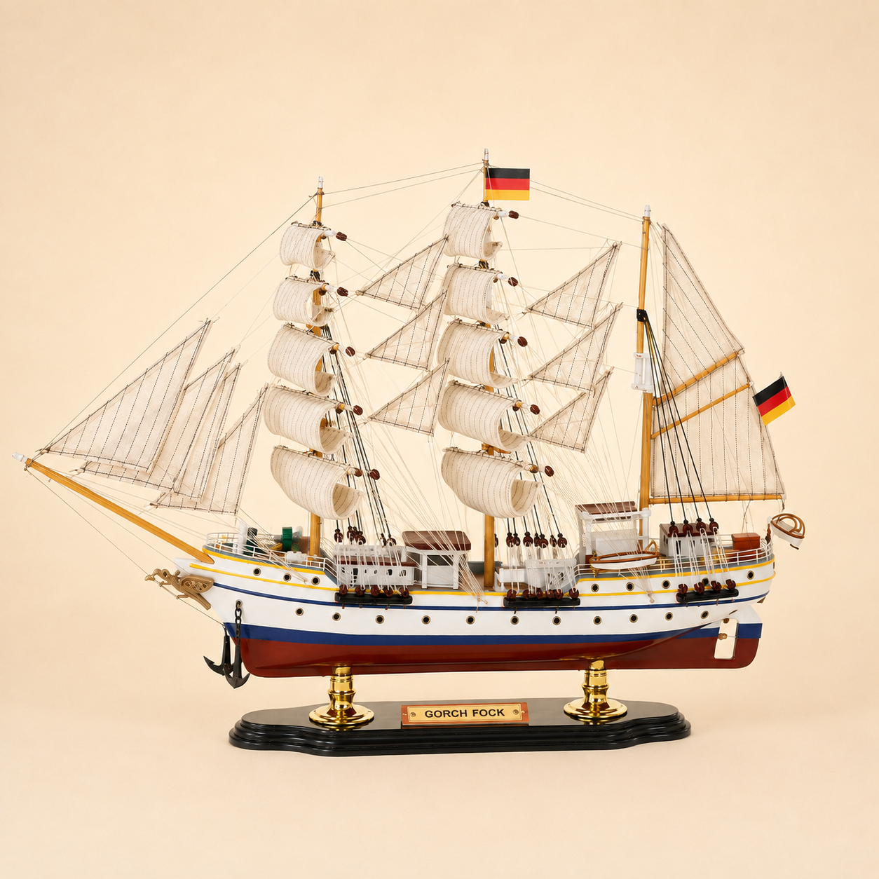 Side view of the Gorch Fock three-masted tall ship model featuring cream sails, intricate rope rigging, white hull, blue and gold trim, red lower hull and black base with gold supports.