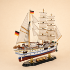 Rear 45-degree view of the Gorch Fock wooden ship model showing the stern, rear deck, German flags, cream sails, detailed rigging, lifeboats and black presentation base.