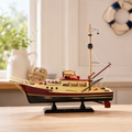 Vintage wooden fishing boat model displayed on a light wood table in a bright nautical interior, featuring a cream and red cabin, black hull, tall mast, working crane, lifebuoy and black display base.