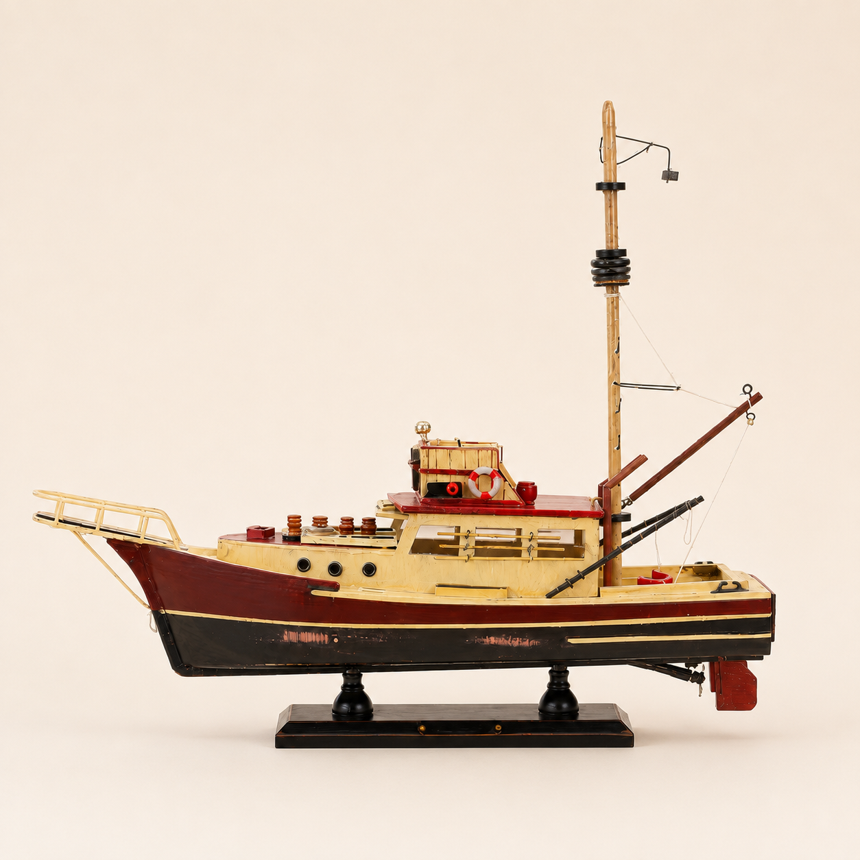 Front 45-degree view of a handcrafted wooden fishing boat model on a cream background, showing the raised bow railing, weathered red and black hull, cream cabin, mast, crane and display stand.