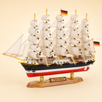 Front 45-degree view of the Preussen wooden sailing ship model on a cream background, showing the bow, five masts, layered cream sails, intricate rope rigging, black anchor, German flags and wooden presentation stand.