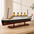 Large RMS Titanic model ship displayed on a light wood table in a bright modern interior, featuring four yellow funnels, detailed lifeboats, black and burgundy hull, Union Jack flag and wooden display stand.