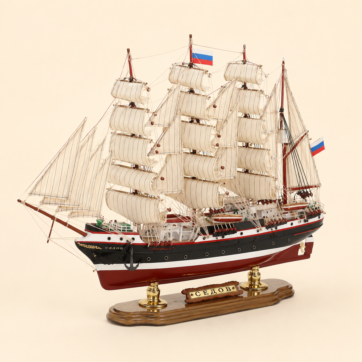 Front 45-degree view of the Sedov wooden sailing ship model on a cream background, showing the bow, layered cream sails, red masts, black anchor, Russian flags and wooden presentation stand.