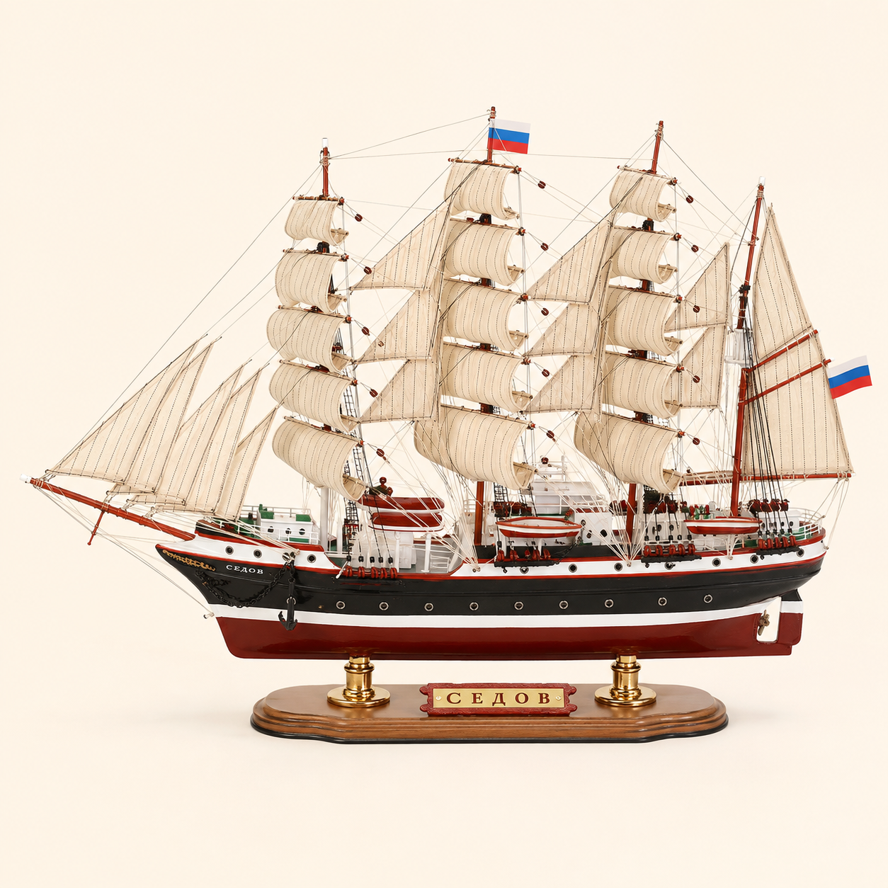 Side view of the Sedov four-masted tall ship model featuring cream fabric sails, intricate rope rigging, black and white hull, red lower hull, lifeboats and a wooden base with gold supports.