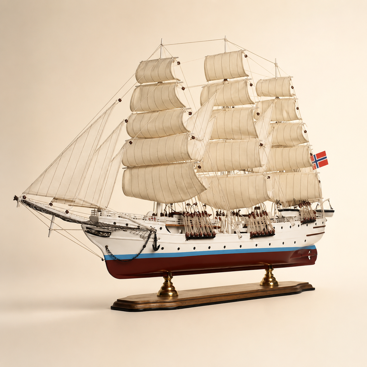 Front 45-degree view of a large Norwegian sailing ship model on a cream background, showing the bow, layered fabric sails, intricate rope rigging, white and red hull and wooden display base.