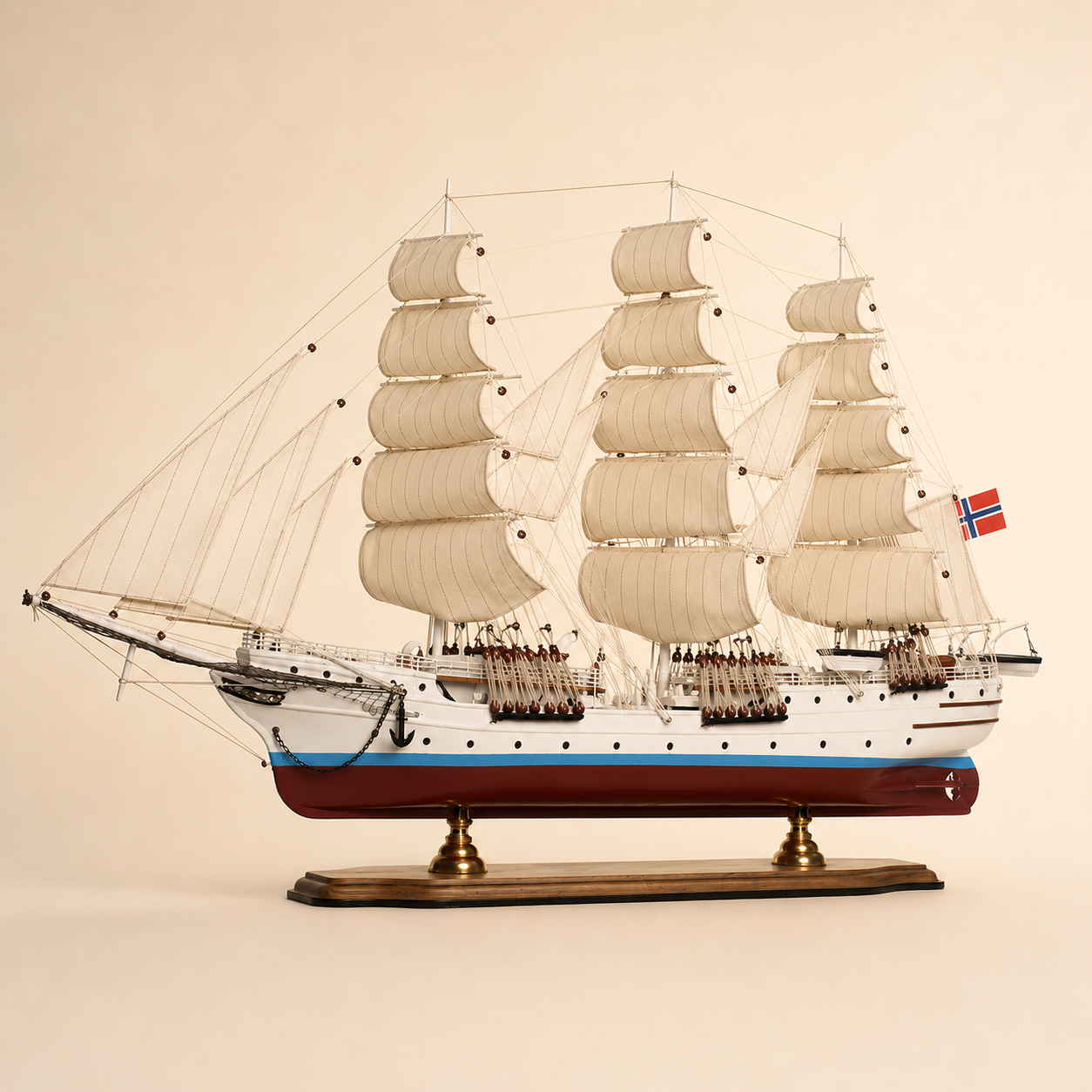 Side view of a large three-masted Norwegian tall ship model featuring cream fabric sails, detailed pulleys and rigging, white hull with blue stripe, red lower hull and wooden stand.