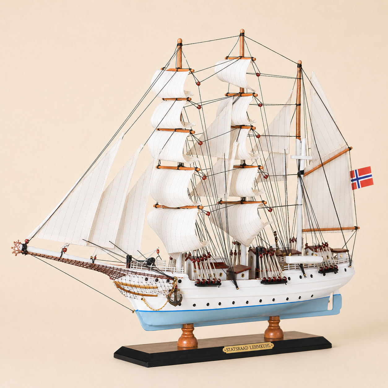 Front 45-degree view of the Statsraad Lehmkuhl ship model on a cream background, showing the bow, layered white fabric sails, detailed rigging, light blue hull and Norwegian flag.
