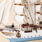 Close-up of the Statsraad Lehmkuhl model ship showing textured white sails, wooden masts, black rigging, pulleys, deck cabins, railings, anchor chain and handcrafted hull details.