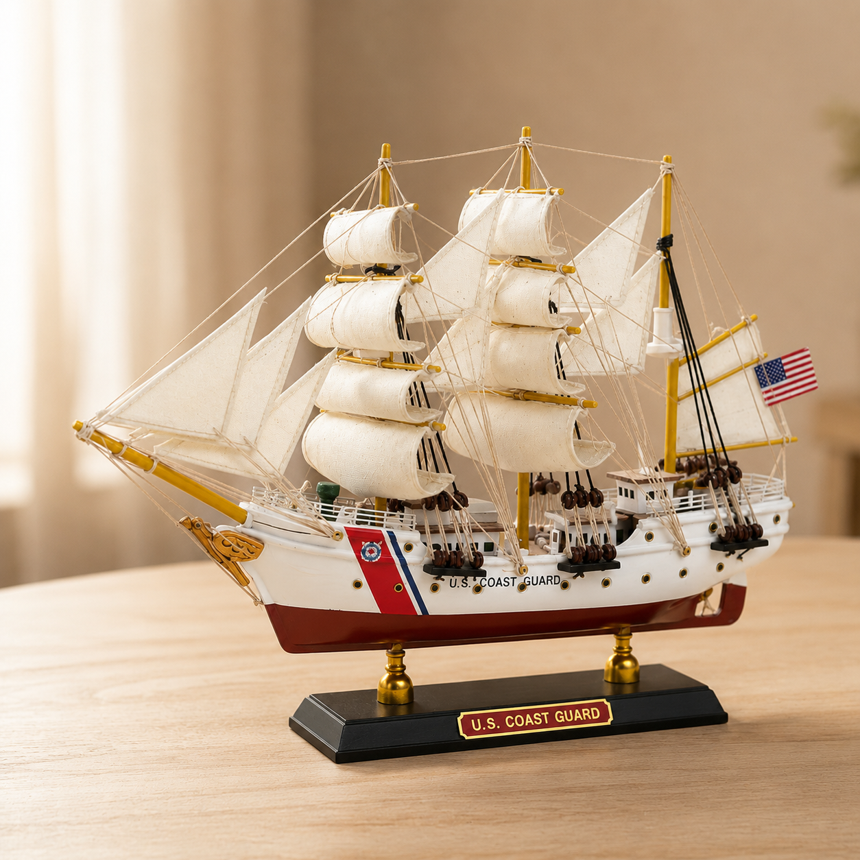 Handcrafted U.S. Coast Guard tall ship model displayed on a wooden table, featuring cream fabric sails, detailed rope rigging, white and red hull, American flag and black display base.