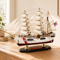 U.S. Coast Guard wooden tall ship model displayed on a light wood cabinet in a bright modern interior, featuring white fabric sails, detailed rigging, American flag, white and burgundy hull, black base and gold supports.
