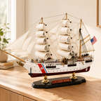 U.S. Coast Guard wooden tall ship model displayed on a light wood cabinet in a bright modern interior, featuring white fabric sails, detailed rigging, American flag, white and burgundy hull, black base and gold supports.