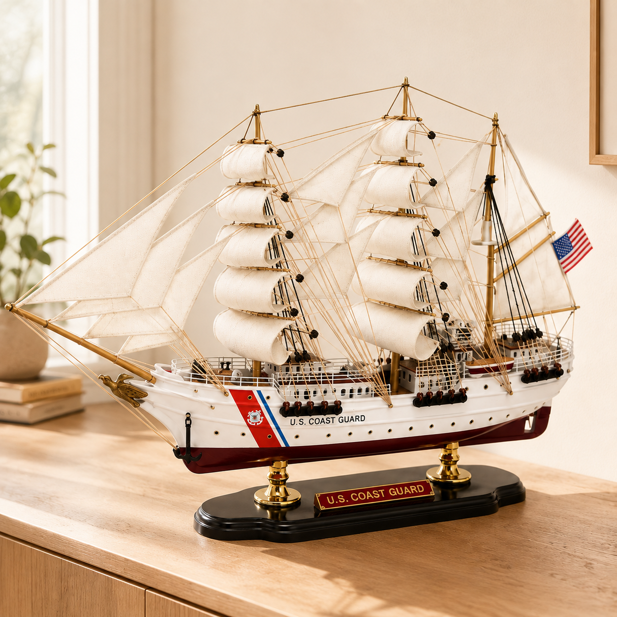 U.S. Coast Guard wooden tall ship model displayed on a light wood cabinet in a bright modern interior, featuring white fabric sails, detailed rigging, American flag, white and burgundy hull, black base and gold supports.