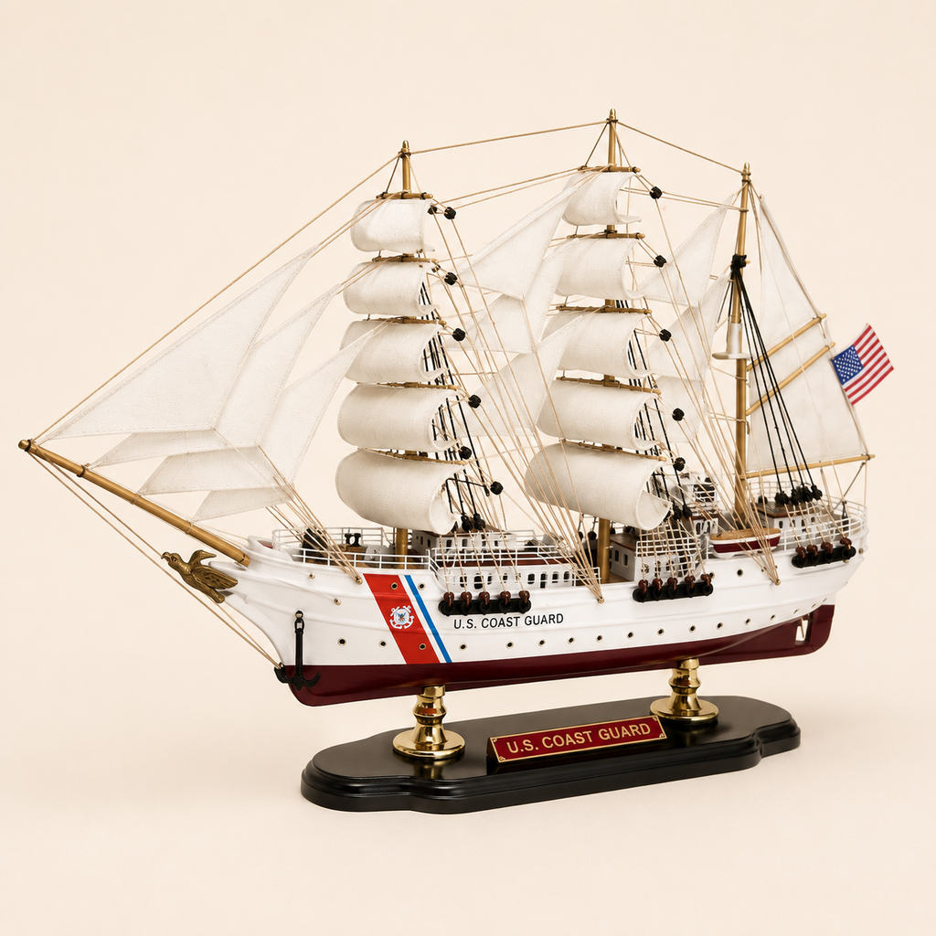Front 45-degree view of the U.S. Coast Guard tall ship model on a cream background, showing the bow, layered white sails, gold-tone masts, black anchor, Coast Guard stripe and American flag.