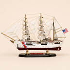 Side view of the U.S. Coast Guard three-masted ship model featuring white fabric sails, detailed rope rigging, white hull, burgundy lower hull, Coast Guard markings and black presentation base.