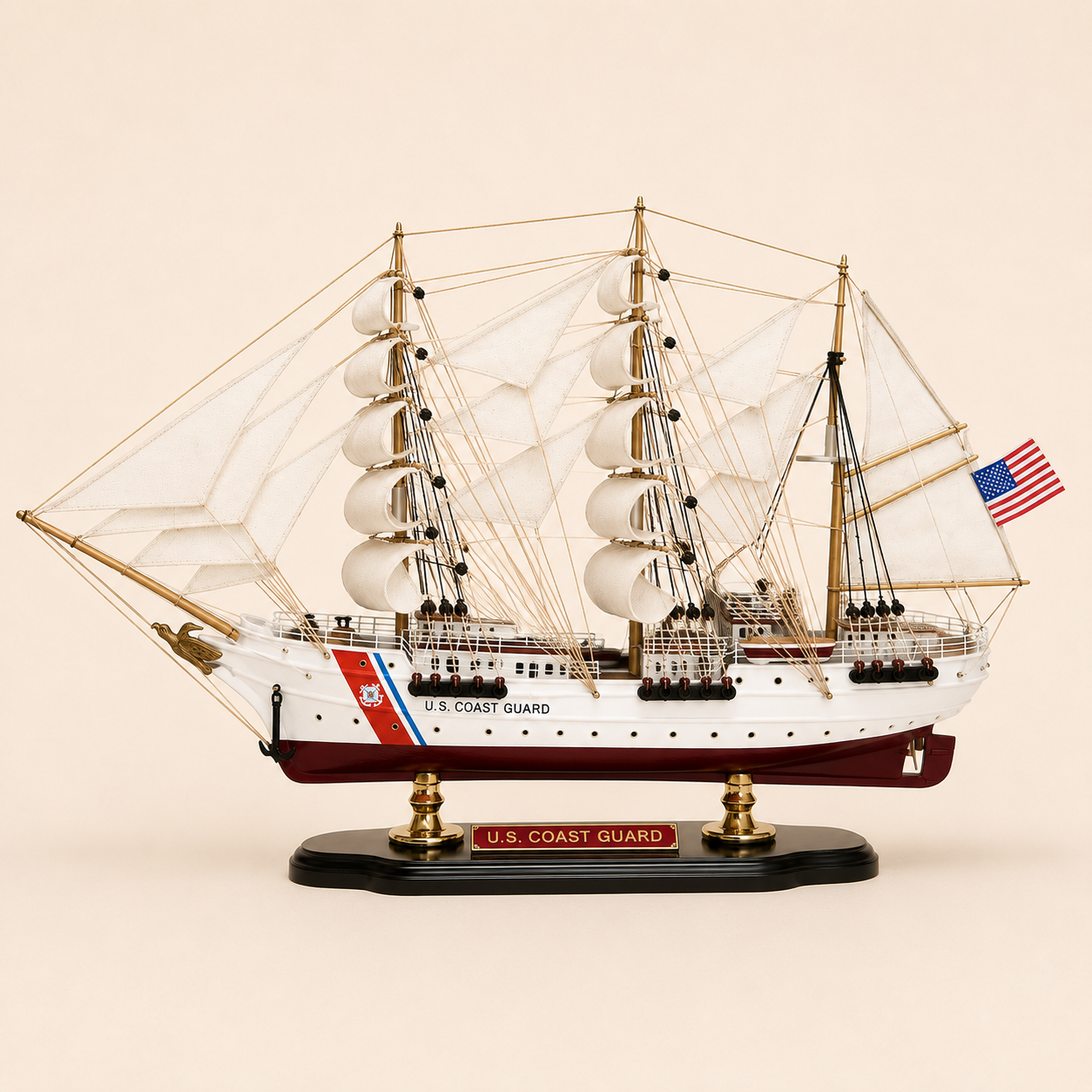 Side view of the U.S. Coast Guard three-masted ship model featuring white fabric sails, detailed rope rigging, white hull, burgundy lower hull, Coast Guard markings and black presentation base.