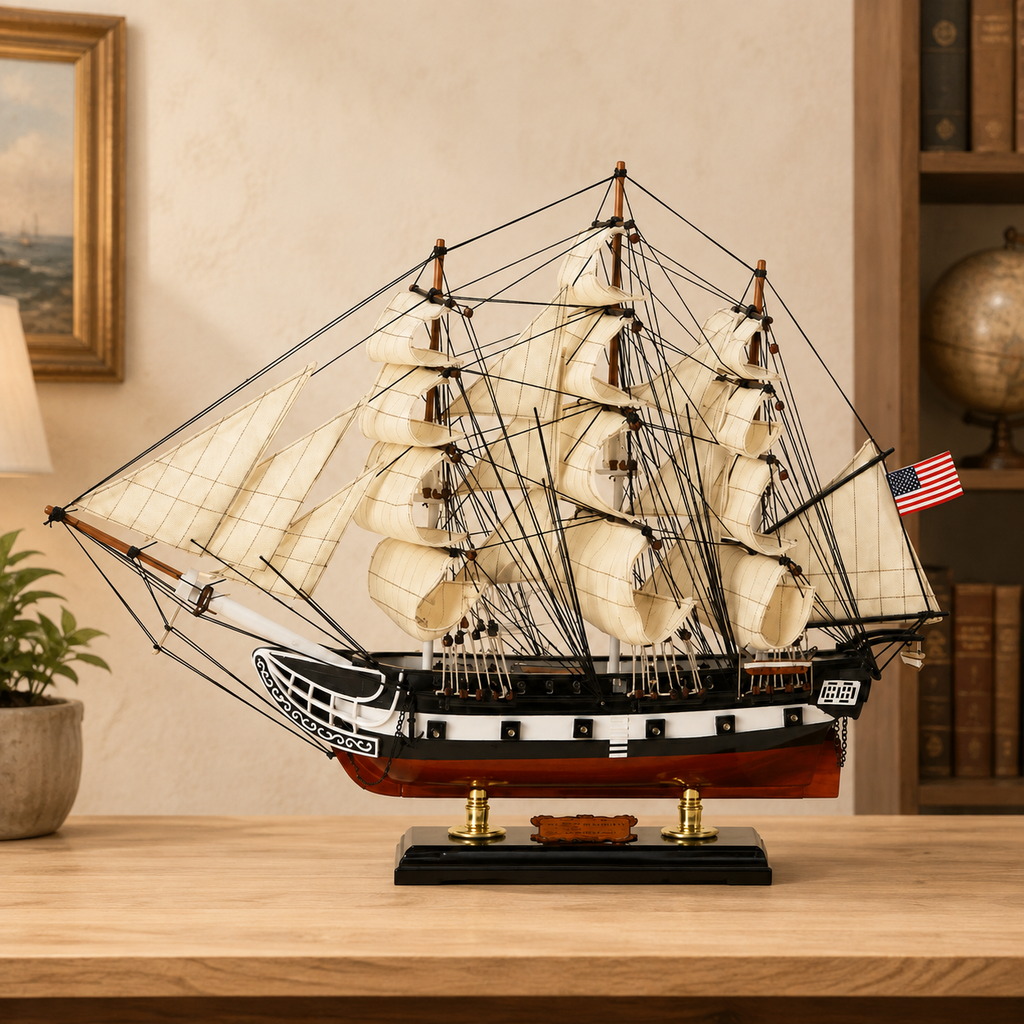American wooden tall ship model displayed on a light wood table in a warm study, featuring cream fabric sails, dense black rigging, a black and white hull, red lower hull, U.S. flag and black display base.