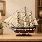 American wooden tall ship model displayed on a light wood table in a warm study, featuring cream fabric sails, dense black rigging, a black and white hull, red lower hull, U.S. flag and black display base.