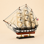 Front 45-degree view of an American wooden tall ship model on a cream background, showing the ornate bow, cream sails, wooden masts, black rigging, U.S. flag and gold-supported display stand.