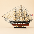 Side view of an American three-masted tall ship model featuring layered cream sails, intricate black rope rigging, black and white hull, red lower hull and polished black presentation base.